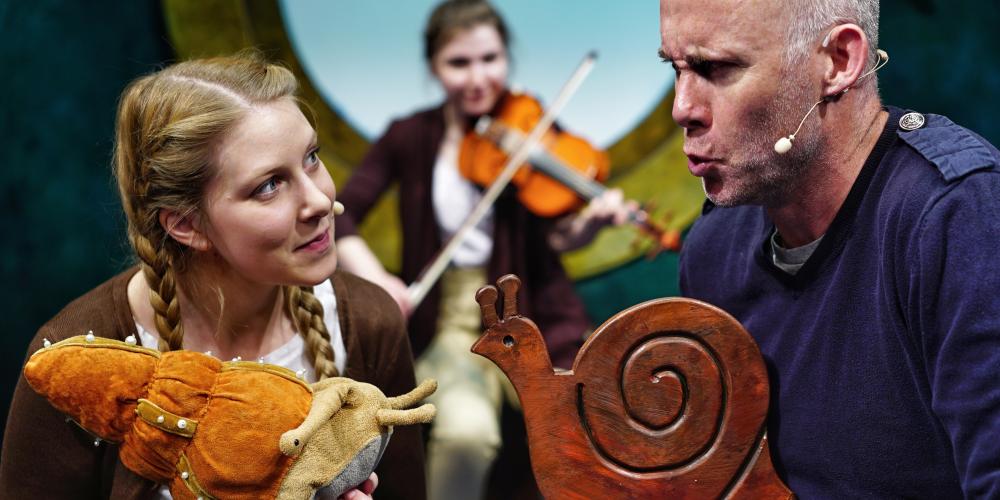 A young woman and an older man both hold small snail sculptures in their hand and talk while another woman plays violin behind them