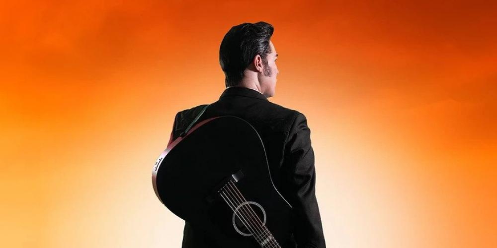 A man in black has his back to us, standing on railroad tracks with a guitar on his shoulder