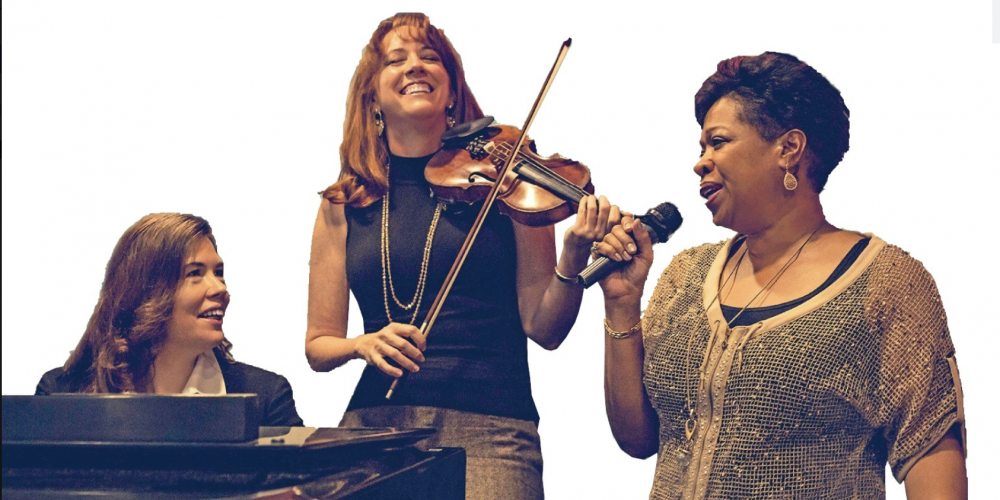 There are three women, smiling - one plays the violin, one plays the piano and the other is singing. 