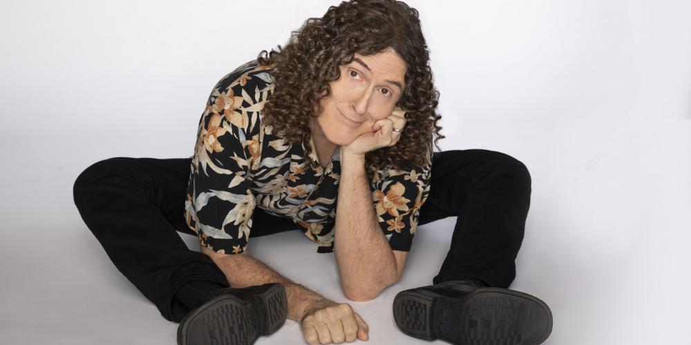 A man with long curly hair, sits on the floor leaning on his elbow