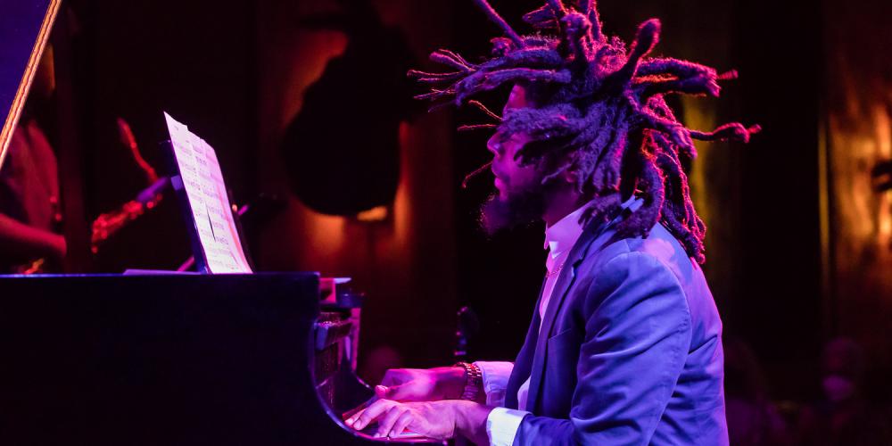 Sean Mason in a suit, playing piano on stage lit with dramatic purple lighting with his dreadlocks standing at attention