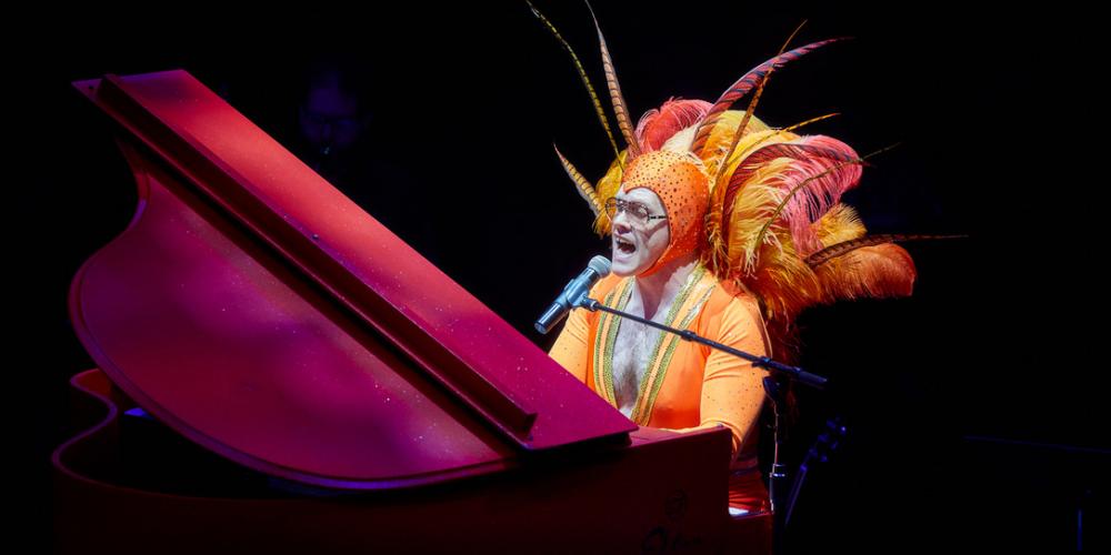 A man sits at a grand piano dressed in an orange spandex outfit and feathered head dress