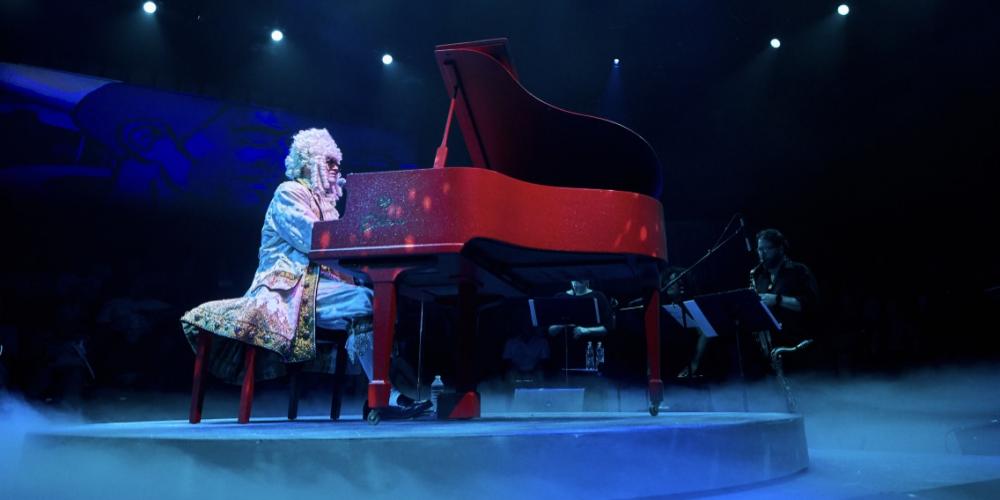 A man sits at a grand piano wearing Louis VIX era clothing and powdered wig