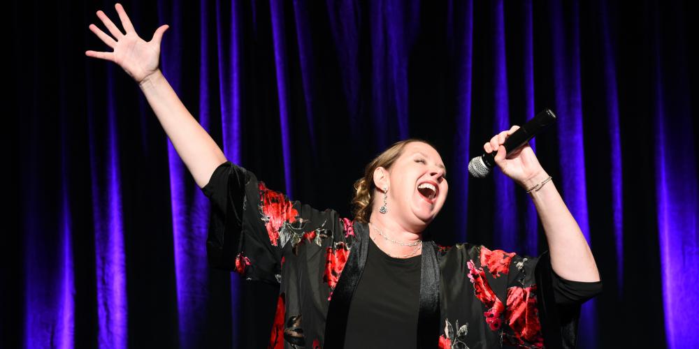 Kristen Drathman sings passionately with one hand raised high in the air and the other holding a microphone to her open mouth.