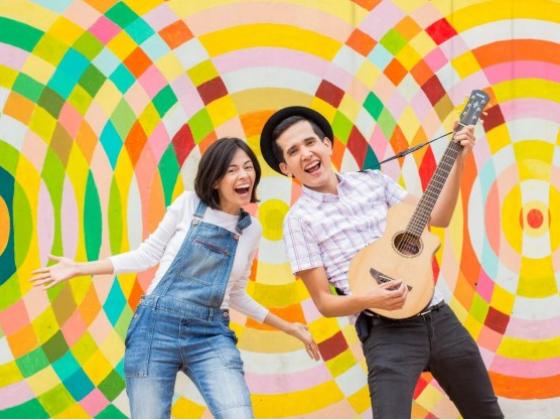A woman dressed in jean overalls and a man wearing a hat, playing guitar, smile at the camera