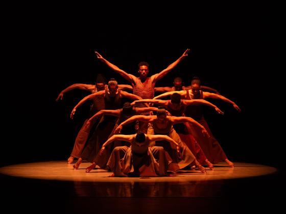 Group of 9 dancers under a spotlight with arms outstretched wide
