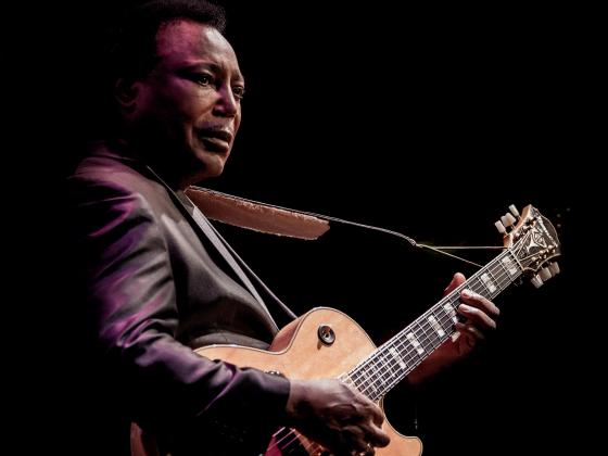 A man with short black hair wears a grey sport coat. He holds an electric guitar, fingers on the fret and strings.