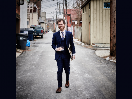 Ben Folds walking on a street in a blue suit and necktie; holding a glass tumbler in one hand and an album in the other. Ben Folds