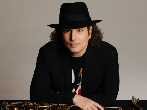 A man with shoulder length curly hair leans over a counter, a saxophone under his hands. He wears all black and a black fedora, smiling slyly at the camera