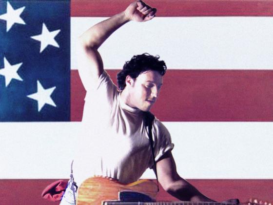 A man wears a white t-shirt with the sleeves rolled up and jeans. He is playing the guitar and leaping with an American flag behind him