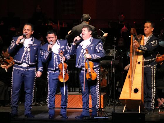 Four men wear blue mariachi suits with silver buckle accents. Two hold violins, one plays a harp and the other sings