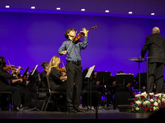 A young man plays the violin on stage with an orchestra behind him