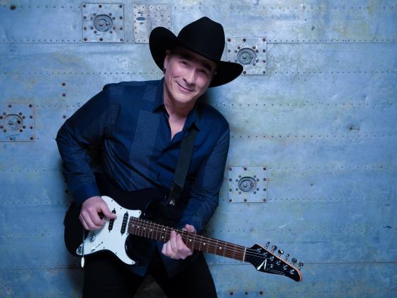 A man in a dark blue shirt stands against a blue wall, electric guitar in hand and a black hat on his head