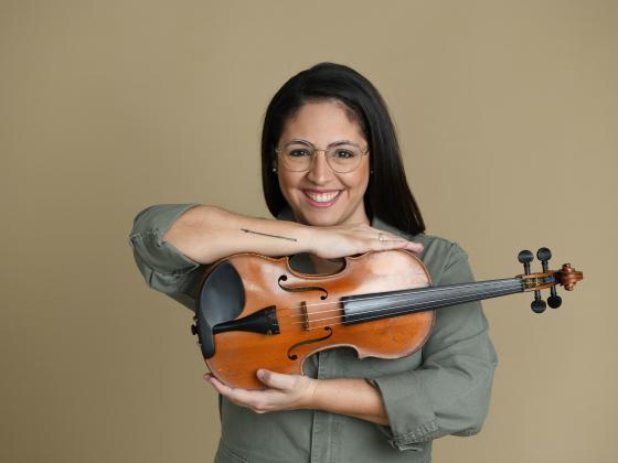 A woman with long black hair and glasses faces the camera holding her violin between her arms