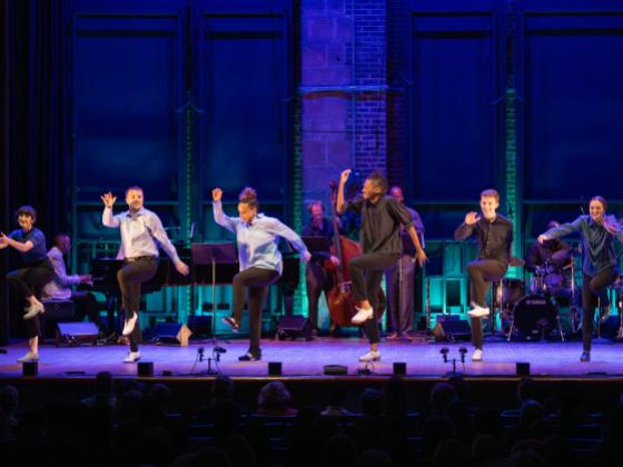 six dancers on stage smiling with right arms up and left knees raised up