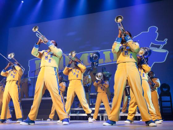 Horn line from Drumline Live wearing yellow uniforms in front of a blue backdrop.