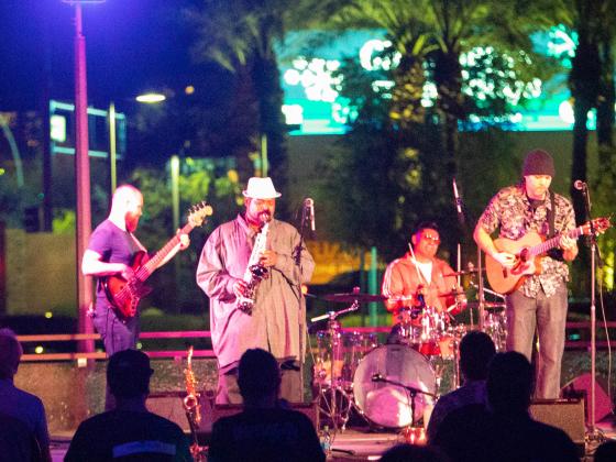 The CCA and Intel present the Free Summer Concert Series with Notes from Neptune Four men play music on a stage that is outside. It is dark out and lights of the city are behind them. Two play guitar, one plays drums and the lead man plays a saxophone.