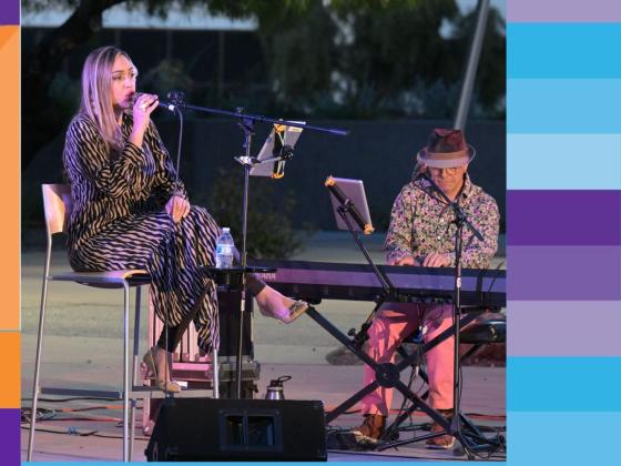 A woman sits on a tall chair on a stage singing into a microphone with a man playing the keyboard to her left