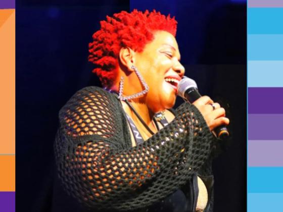 A woman with short red curly hair, black shirt and large gold earrings sings into a microphone