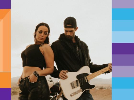 A woman wearing black stands close to a man who is wearing black and holding a white electric guitar