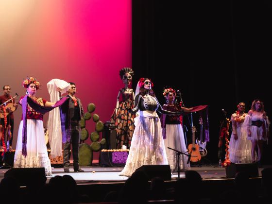 8 performers dressed in dia de los muertos themed attire  on stage with pink backlight 