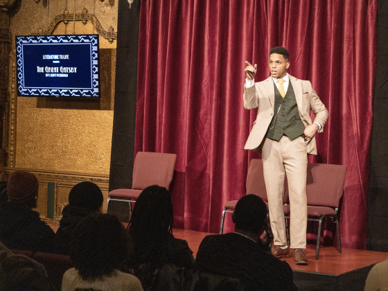 A man wearing a brown suit and green vest stands on a stage talking to the audience
