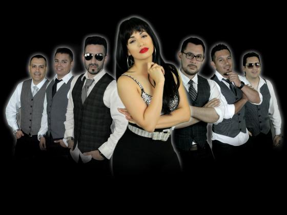 A woman with long black hair and bright red lips, wears all black with a silver studded top. Her hand is under her chin and her hip juts out to one side. Six men, three on either side of her, wear white button up shirts, black ties and vests
