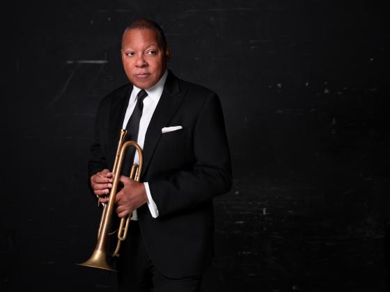 a man in a black suit stands against a black wall with a trumpet