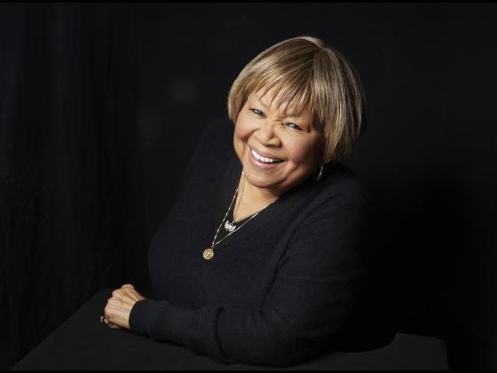 Mavis headshot with her smiling at the camera leaning on one arm and dressed in black
