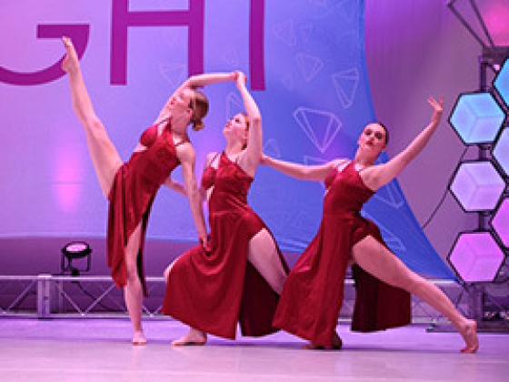 three dancers with arms and legs outstretched on stage in red dresses
