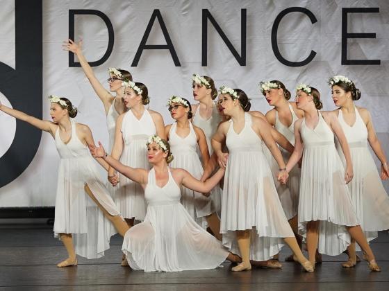 A group of barefoot modern dancers dressed in long white dresses pose on a stage