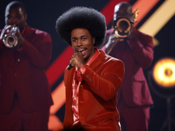 singer dressed in red singing on stage with trumpeters in background