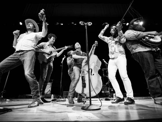 Four men dressed in jeans and traditional western wear hold string instruments while performing on stage
