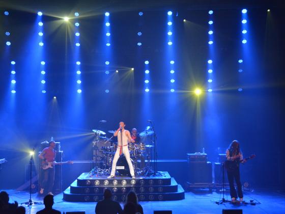 A man dressed in white jeans and a white sleeveless shirt stands center stage with blue lights streaming behind him