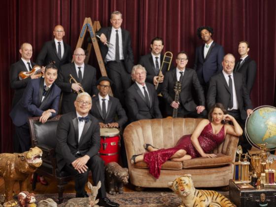 Thirteen men wearing black suites and a women in a red dress laying on her side on a sofa. 