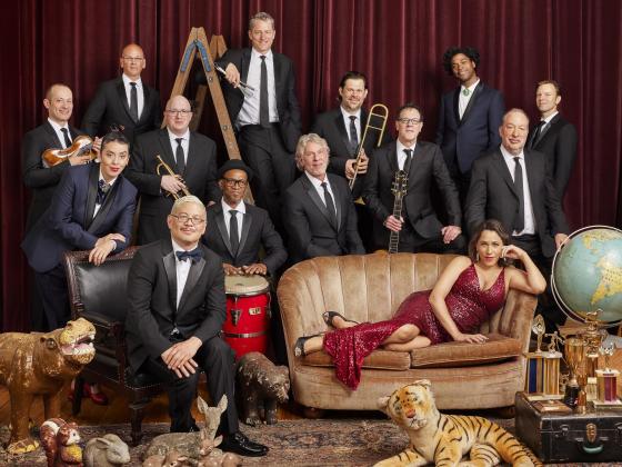 14 people dressed in formal attire - 13 in tuxedos and one lounging in a red sequin dress on a couch