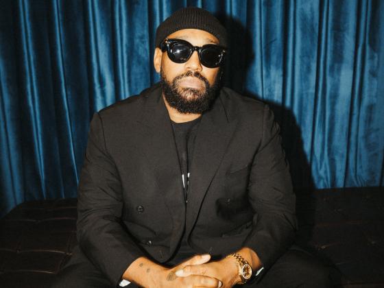 A man wears a black shirt, black suit jacket, black beanie hat and black sunglasses, facing the camera 