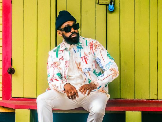A man sits on a red bench wearing white pants, a white tropical print button up shirt, sunglasses and a black beanie
