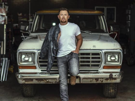 Scotty McCreery leaning back on front of an old truck wearing jeans and t-shirt