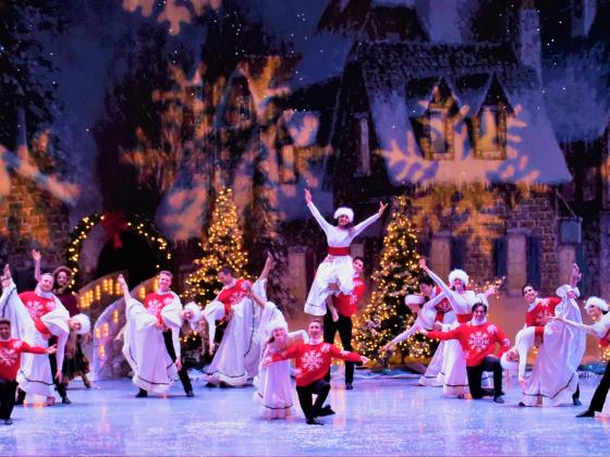 Spirit of Christmas Performance Kickline Performers dressed in white and red posing and smiling on stage in front of a glowing snowflake scene