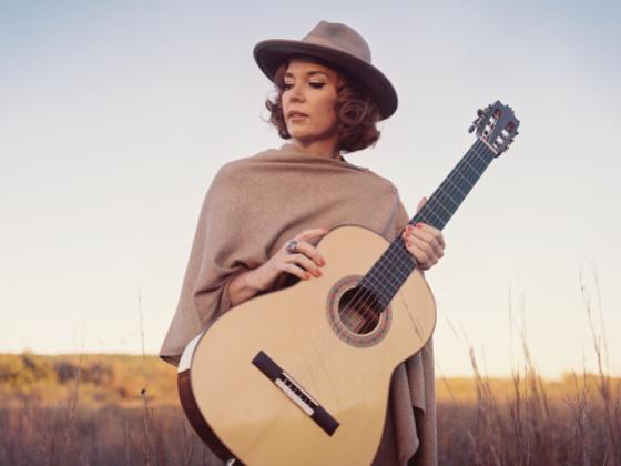 Sue Foley in field of grass looking off to the side in a hat and shall holding an acoustic guitar
