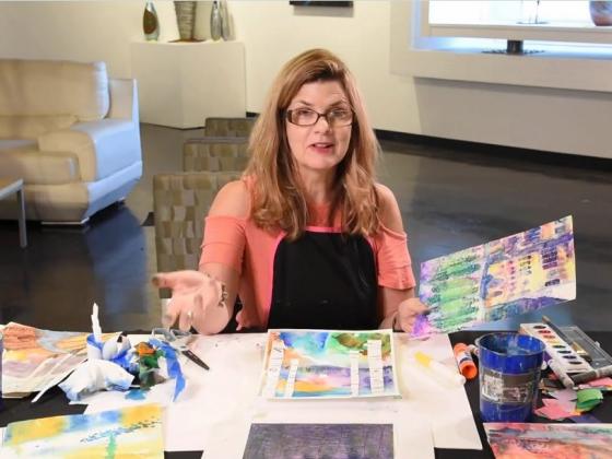 CCA Anywhere presents Watercolor Wash with Deb Goley A female wearing glasses and a coral colored shirt sits at a table with art supplies and paper laid out on it