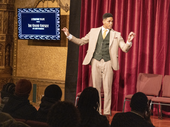 A man wearing a tan suit with a green vest talks to an audience from a stage