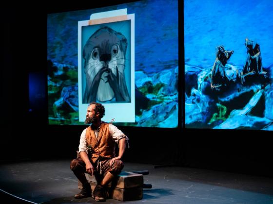 Theatre Kids: Wind in the Willows Man sitting on stage with a photo of otter on a projected screen