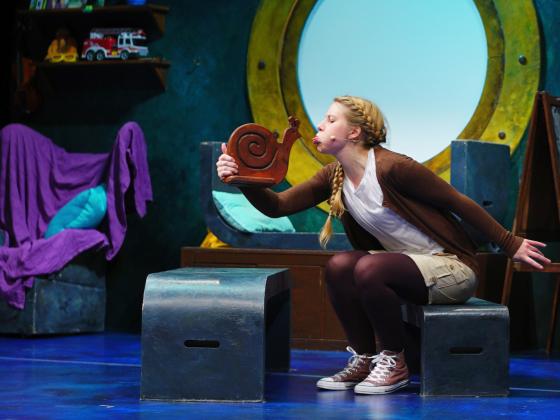 Virtual Theatre Kids: The Snail and the Whale An image of a young woman sitting in a room that looks nautical, talking to a small snail sculpture in her hand