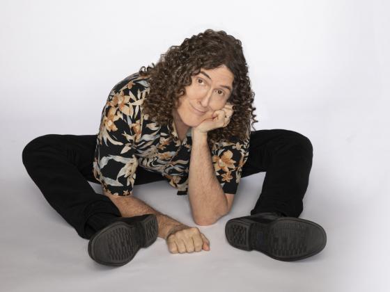 A man with long curly hair, sits on the floor leaning on his elbow by his feet. He is  wearing black pants and a patterned shirt