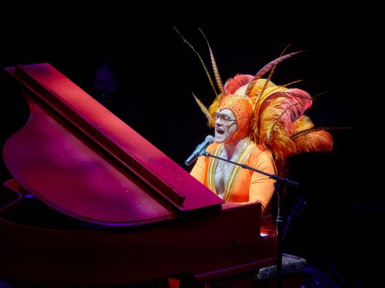 Chandler Center for the Arts presents Wonderful Crazy Night: An Elton John Tribute A man sits at a grand piano dressed in an orange spandex outfit and feathered head dress