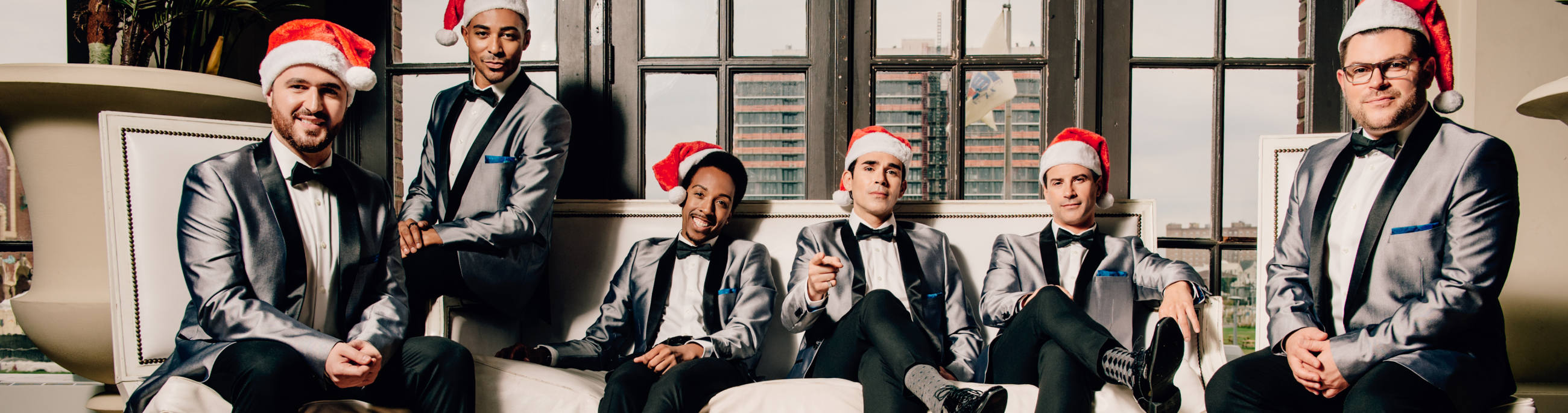 Six men dressed in black and gray suits and wearing santa hats, sitting on a couch looking at the camera