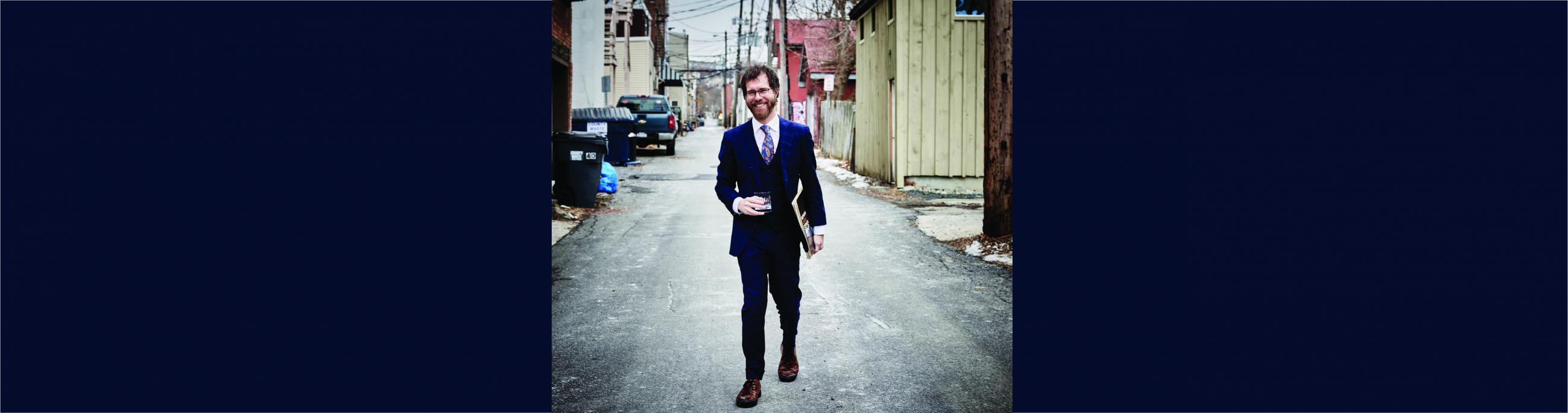 A man with glasses, a beard and brown hair wears a suit, walking in an alley.