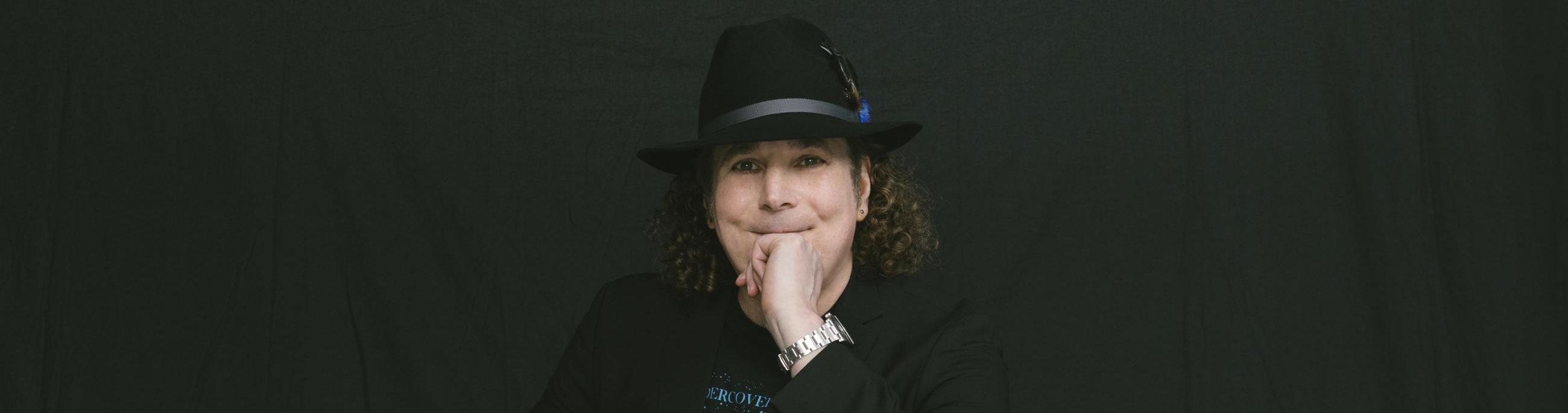 white man with curly brown hair wearing all black and a black hat in front of a black background.